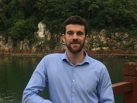 man in a blue shirt smiling standing in front of a lake