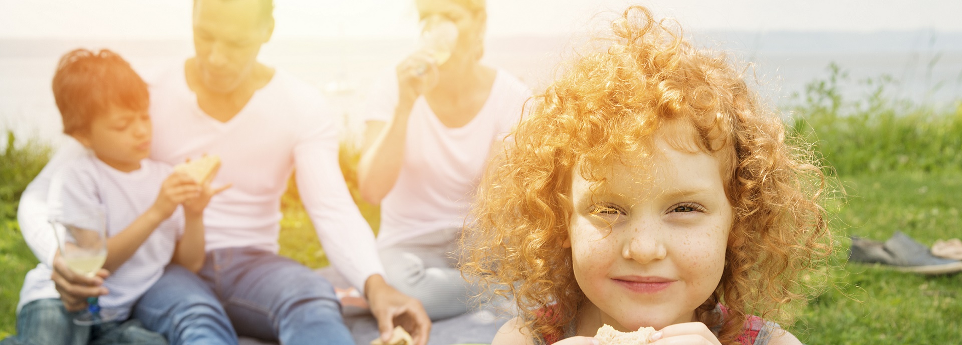 Girl with red, curly hair eating a sandwich with man and woman in the background