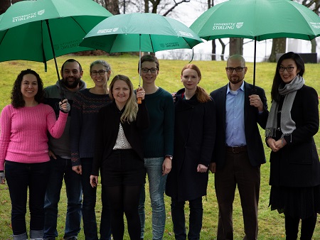 Group of eight people standing under three University umbrellas