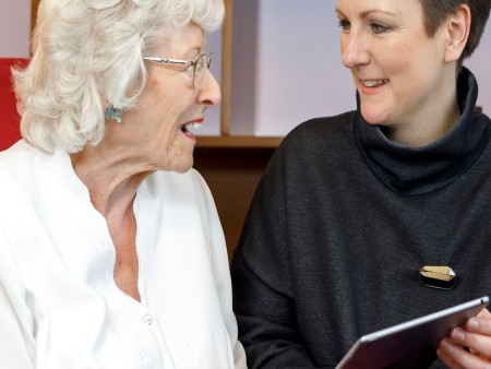 Two women sat next to each other. The younger one is showing the other elderly lady something in a book. 