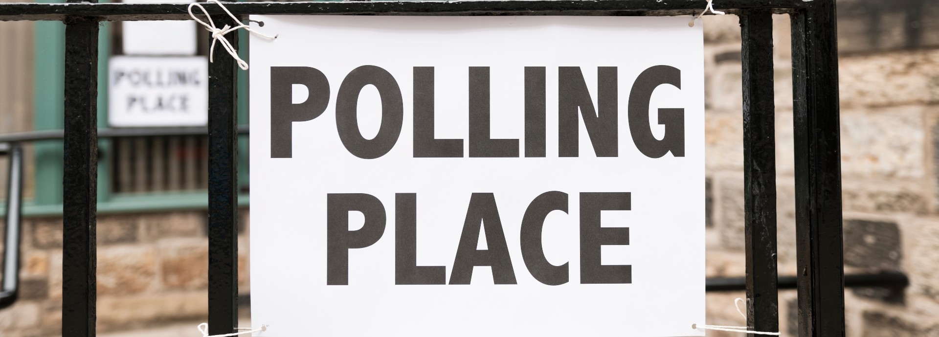 A sign for a polling station tied to a black metal fence with a brick building in the background