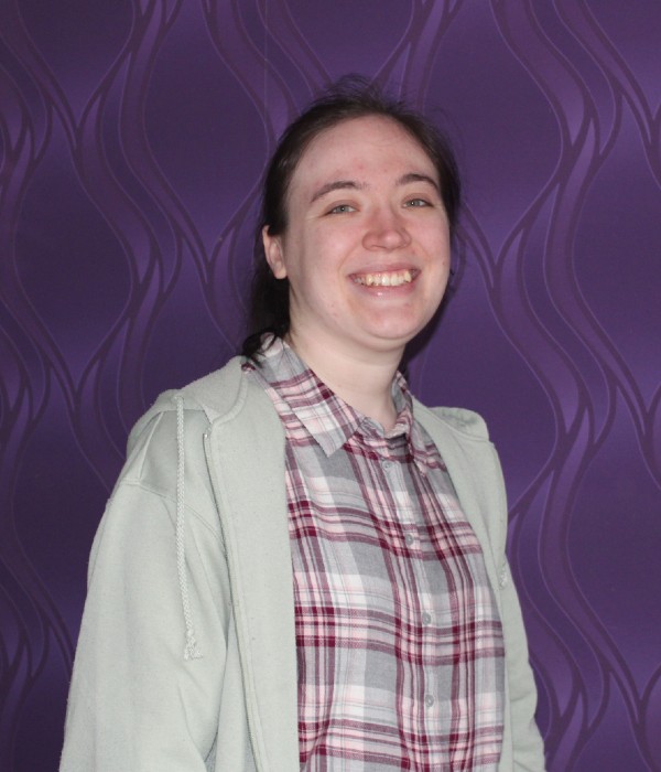 Jacque, a 23-year-old female wearing a red and white checked shirt and cream cardigan. She has dark hair swept off her face and is smiling directly to camera.