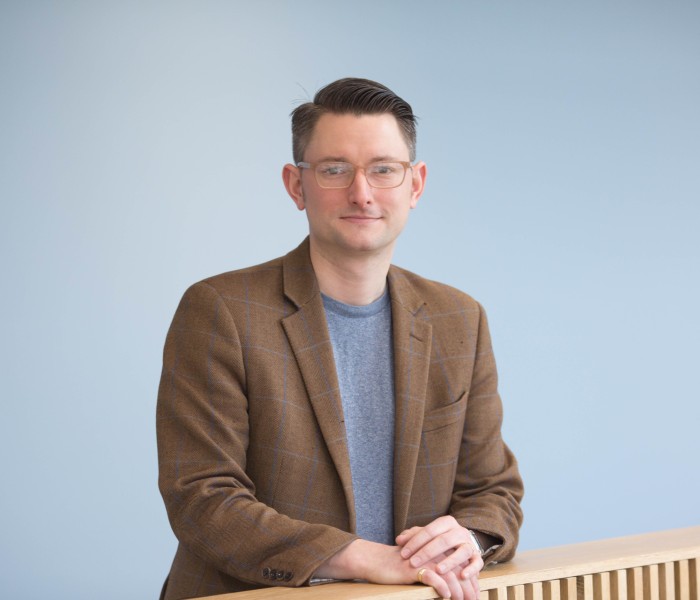 Dr Stuart Auld, a man with short dark hair wearing small, square glasses, a brown blazer and a grey t-shirt. He looks straight to camera.