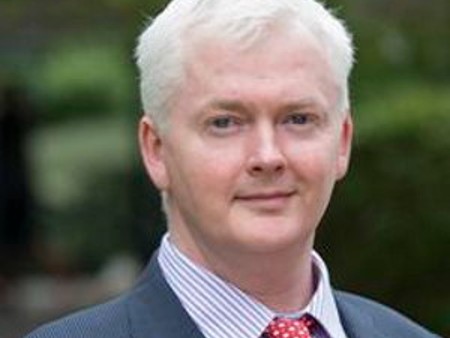 Photograph shows Prof Derek McGhee. It is a head and shoulders shot, with Prof McGhee pictured outside and looking straight to camera. He has white hair and is wearing a grey suit, light coloured shirt and red tie