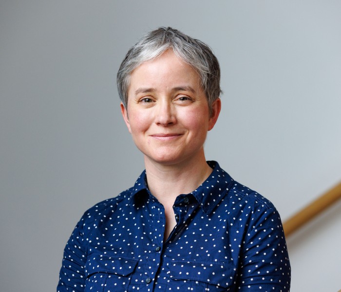 Portrait photograph of Kirstie Blair. She has short grey hair and is wearing a blue polka dot blouse. She is pictured indoors, against a grey background.