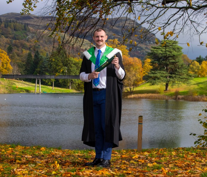 Ross poses next to loch in graduation gown holding medals