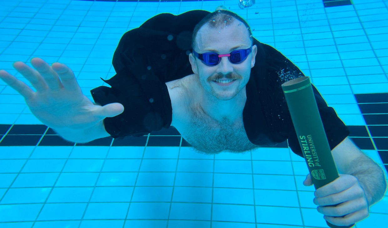 Ross in the swimming pool wearing his graduation gown and clutching a University scroll underwater