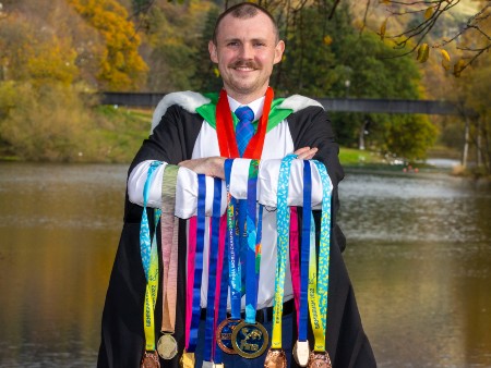 Ross poses next to loch in graduation gown holding medals