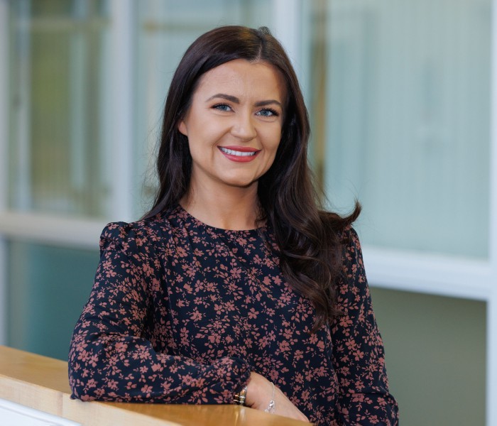 Amy McCarron leans on a bannister. She has long, dark hair and is weather a floral patterned top.