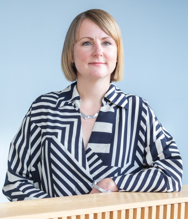 A portrait photograph showing a woman pictured inside against a blue background and leaning on a light coloured wood bannister. She is wearing a black and white stiped shirt, silver necklace and has short light brown hair.