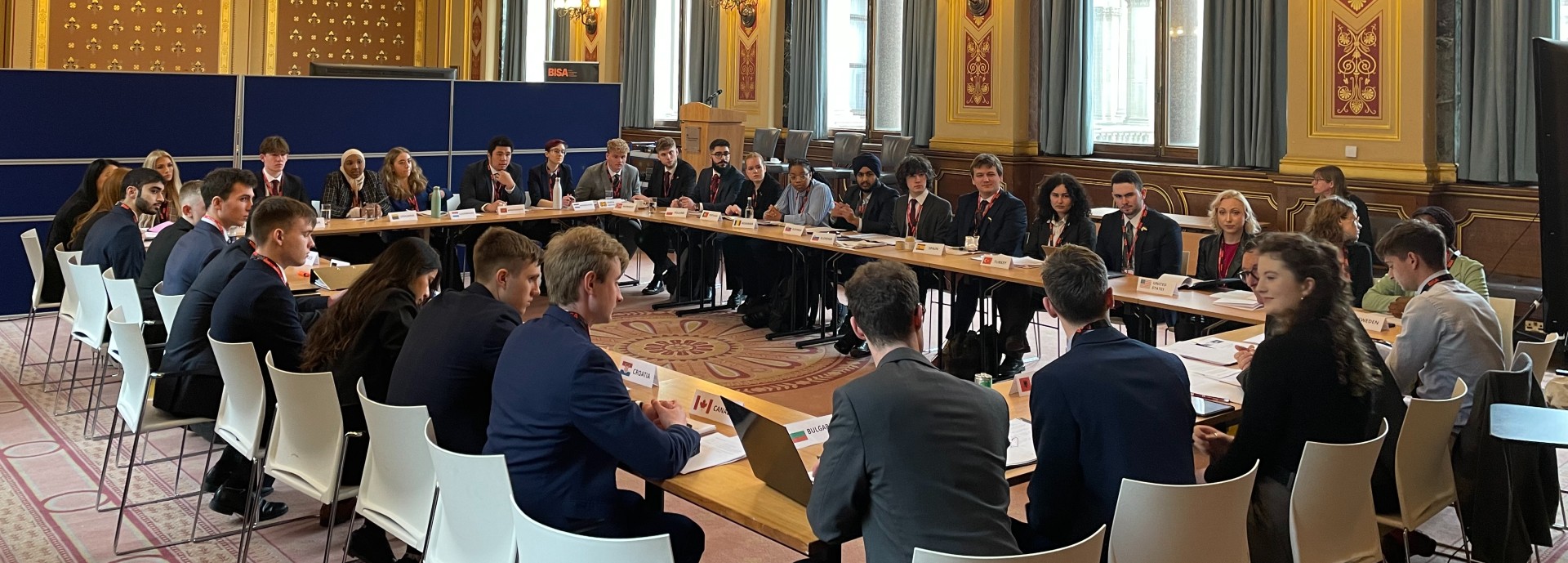 Photograph shows students taking part in 'Model NATO' sat round a large table in the offices of the Foreign Office, London