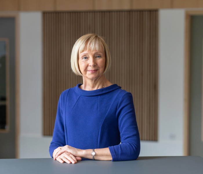 Prof Karen Watchman poses with her arms crossed. She has blonde bobbed hair and is wearing a blue dress.