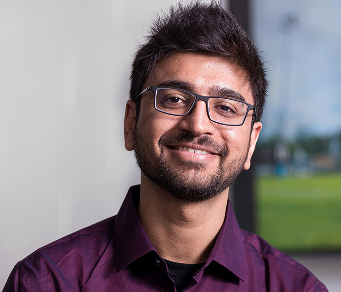 Male Indian student wearing glasses and purple shirt standing in front of a screen displaying football image