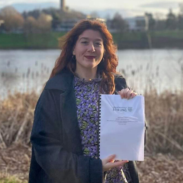 Billi is stood near the loch on campus holding their dissertation